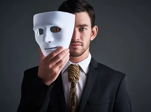 Uomo in abito elegante tiene in mano uno maschera bianca, parzialmente sollevata davanti al viso. Lo sfondo è scuro e l'atmosfera è intensa e drammatica.