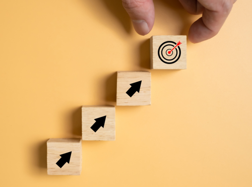 Blocchi di legno disposti a gradini con frecce verso l’alto e un bersaglio finale, simbolo di un percorso di crescita e obiettivi da raggiungere.