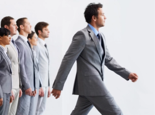 Gruppo di professionisti in abito grigio allineati; uno di loro avanza deciso davanti agli altri, che restano ferm