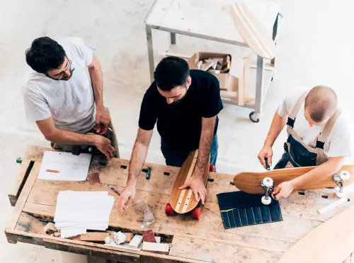 Tre artigiani di generazioni diverse lavorano insieme su un banco da falegname: il mestiere si impara affiancando chi lo pratica.